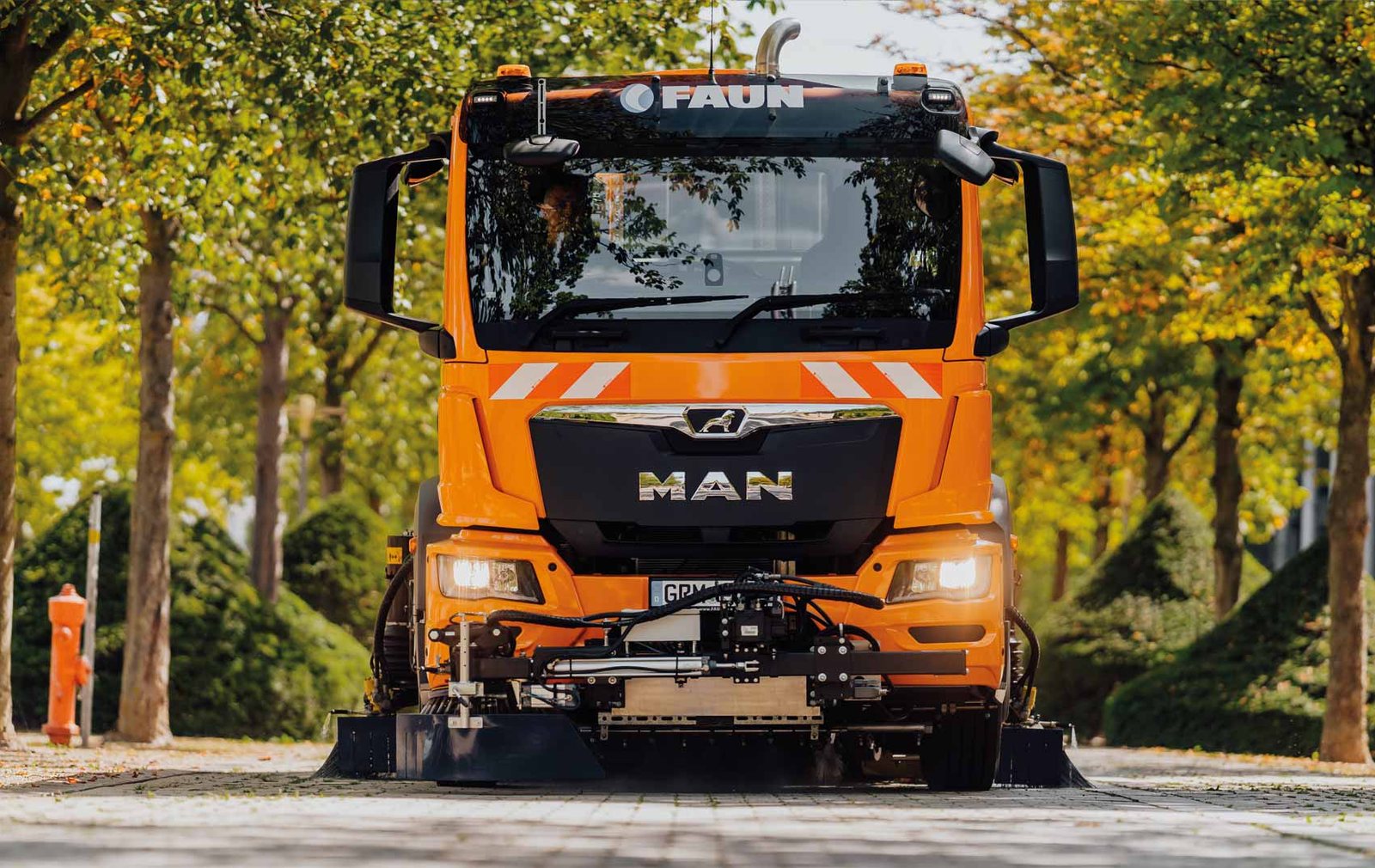Orangefarbene FAUN Viajet Kehrmaschine von vorn, zwischen Bäumen