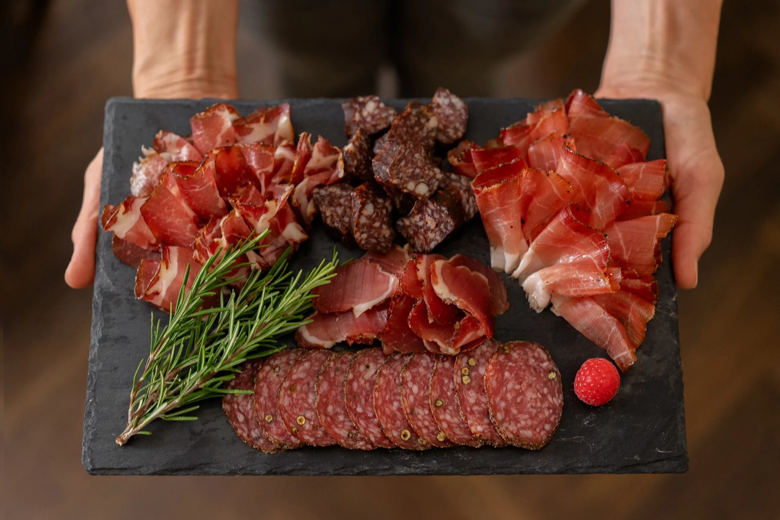 Hände halten eine Schieferplatte mit Schinken- und Wurstspezialitäten von Tiroler Delikatessen